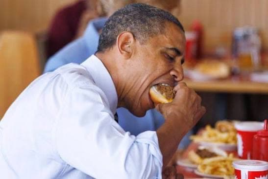 A man in shirtsleeves taking a large bite of a cheeseburger in a diner booth